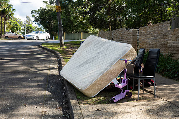 A large pile of rubbish set out on the curb for council collection in a suburban neighbourhood. The heap includes oversized items like flattened cardboard boxes, a worn chair with pillows, mattresses stacked atop one another and other household waste