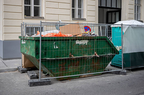 Green dumpster full of construction waste behind metal fence on city street